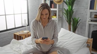 Parlak yatak odasındaki rahat bir yatakta selfie çeken bir kadın. Yumuşak ışıklandırma ve doğal elementler içeren modern dekorasyonla huzurlu bir atmosferi geliştiriyor..