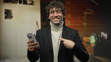İnşaat alanında, balıkçı yaka ve ceket giymiş, kentsel tarzı ve modern iletişimi yansıtan akıllı bir telefonla iletişim kurarken kapalı alanda gülümseyen genç bir adam..