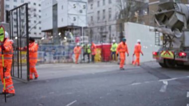 Bir inşaat sahasında turuncu üniformalı ve miğferli işçiler, kentsel zemin ve ekipmanlar arasında bulanık bir odak noktasına sahip, endüstriyel açık hava ortamı ve takım çalışmasını ön plana çıkarıyor..