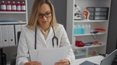 Hastane odasında belgeler okuyan, gözlük takan, laboratuvar önlüğü giyen, arka planda tıbbi ekipman ve malzemeler bulunan bir kadın, profesyonel bir sağlık hizmetleri ortamını ön plana çıkarıyor..