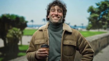 Yakışıklı bir adam yeşil ve okyanus manzaralı manzaralı şehir caddesinde elinde kahve fincanı tutarak gülümsüyor..