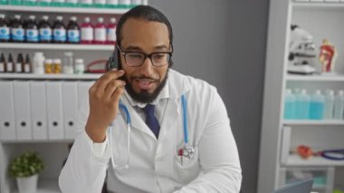 Klinikte sakallı bir adam konuşuyor. Laboratuvar önlüğü giyiyor. Etrafı tıbbi malzemeler ve laptoplarla çevrili..