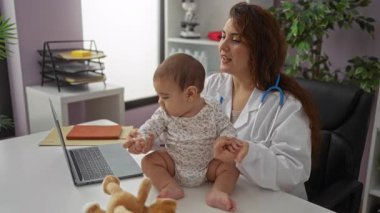 Beyaz önlüklü bir kadın klinikte kucağında laptopuyla ve oyuncaklarıyla bir bebek tutuyor. Kapalı alanlarda sağlık hizmetleri ortamı yaratıyor..