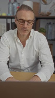 Orta yaşlı, beyaz, sarı saçlı, ofis ortamında masa başında çalışan, gözlüklü, beyaz gömlekli, iç mekan bitkileri ve raflarla çevrili bir adam..