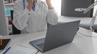 Diz üstü bilgisayar üzerinde çalışan kadın doktor, modern klinik ofisinde, steteskop boynunda, ekrana odaklanmış; organize masa, profesyonel çevre, özenli duruş görünür.