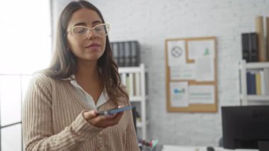 Ofiste telefonla konuşan gözlüklü ve bej kazaklı bir kadın masası ve arka planda rafları olan parlak, modern bir iç mekanda güvenle gülümsüyor..