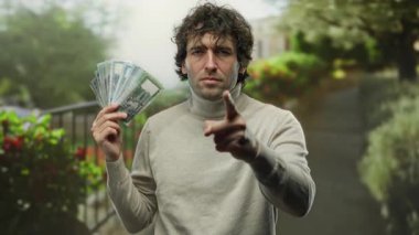 Bangladeş banknotları tutan İspanyol bir adam şehir sokağında kameraya ciddi bir ifadeyle bakıyor, şehir yeşiliyle çevrili..