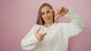Elleriyle çerçeve oluşturan genç bir kadın, süveter giyen mutlu fotoğrafçılık konseptiyle gülümseyen, izole edilmiş pembe arka planda odaklanmış, yaratıcılık ve neşe gösteren bir kadın.