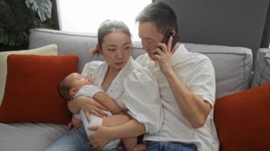 Aile, oturma odasındaki rahat bir kanepede birlikte otururken, anne ve baba telefonda, samimi bir evde sevgi ve ebeveynlik sergiliyorlar..