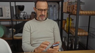 Modern ofis ortamında gözlüklü olgun bir adam raflar ve mutfak gereçleri ile çevrili akıllı telefonu kullanıyor, odaklanmış ve dalgın görünüyor..