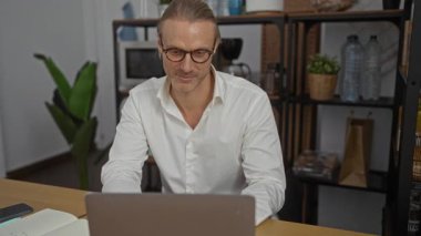 Beyaz gömlekli gülümseyen adam dizüstü bilgisayarda çalışıyor. Etrafı modern çalışma alanlarını gösteren raf eşyaları ve bitkilerle çevrili..