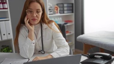 Klinik odasında kızıl saçlı kadın doktor, tıbbi dosya ve ekipman raflarıyla çevrili modern hastane ortamında dizüstü bilgisayarla ve steteskopla çalışıyor..