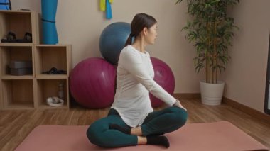 Spor salonunda yoga yapan bir kadın. Etrafı spor topları ve ekipmanlarla çevrili. Pembe bir paspasın üzerinde esneme sırasında rahatlama ve spor yapmayı teşvik ediyor..