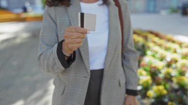 Şehir parkında açık havada kredi kartı dağıtan, spor ceket giyen, canlı çiçekler takan, arka plan bulanıklaşması yaşayan, gündüz vakti görülebilen, ışık odaklı, olgun bir kadın..