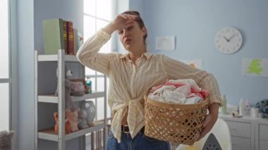Çamaşır sepetini parlak bir iç mekânda tutan kadın, ev içi dekorasyonunu gösteren oyuncaklar ve kitaplarla dolu düzenli bir odada dururken yorgun bir ifade sergiliyor..
