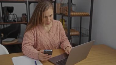 Dizüstü bilgisayarda yazan ve rahat ofis çalışma alanında rafları ve ahşap masası olan akıllı telefonu kullanan sıradan bir kazak giyen, modern çalışma ortamını gözler önüne seren bir kadın..