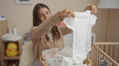 Kadın, oyuncak ve beşikle çevrili rahat yatak odasında bebek kıyafetlerini katlıyor. Sıcak ve sade bir ortam yaratıyor..