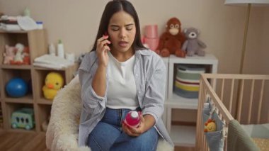Bebek odasında oturan kadın, oyuncak ve bebek malzemeleriyle çevrili bir şişeyi incelerken cep telefonuyla konuşuyor, bakım ve ev ortamını ifade ediyor..