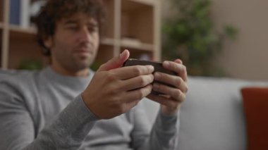 Genç adam kapalı bir oturma odası ortamında akıllı telefonuyla oynuyor, ekranda dikkatle odaklanıyor ve mobil oyun oynarken heyecan ve konsantrasyonunu ifade ediyor..
