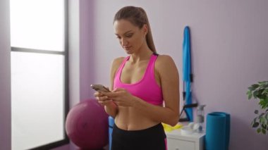 Pembe spor kıyafetli bir kadın spor salonunda telefon kullanıyor. Heyecan ve odak noktalarını içeride gösteriyor. Aktif yaşam tarzı bağlılığını ve dijital bağlılığı gözler önüne seriyor.