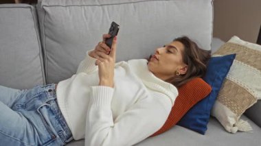 Kanepede uzanmış esmer saçlı, rahat bir oturma odasında akıllı telefon kullanan, rahat ve rahat bir yaşam tarzı sergileyen genç bir kadın..
