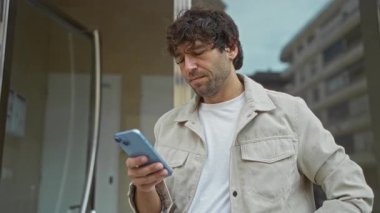 Kentsel sokak ortamında akıllı telefona bakan genç İspanyol bir adam modern yaşam ve teknoloji etkileşimini gösteren düşünceli bir ifadeyle
