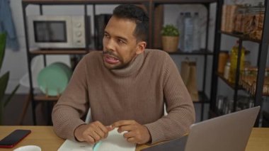 Ofiste çalışan bir adam, dizüstü bilgisayarında masasında otururken düşünceli bir ifadeyi not defterine yazarken gösteriyor.