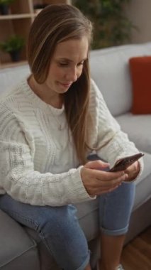 Beyaz kazaklı kadın rahat oturma odasında otururken akıllı telefon kullanıyor. Kanepesi ve çiçekleri gün ışığında şık bir ev içinde görünüyor..