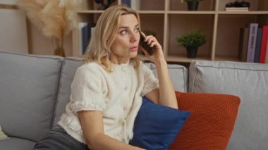 Konforlu oturma odasında rahat bir ev ortamı yayarak, yastıklar ve raflarla çevrili, sürpriz duygular gösteren bir kadın telefonda konuşuyor..