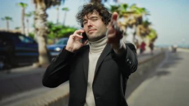 Şehir merkezinde palmiye ağaçlarıyla dolu bir sokakta telefonla konuşurken dışarıda kamerayı işaret eden genç adam..