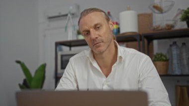 Ofis ortamında çalışan orta yaşlı, beyaz bir adam, profesyonel bir ortamda gözlük tutuyor ve arkasında raflar ve bitkiler var..