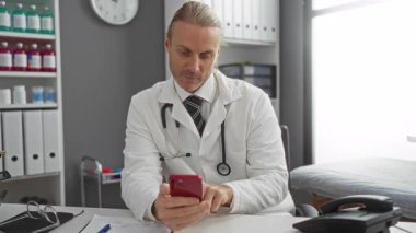 Masasında tıbbi malzemeler ve belgelerle hastane ofisinde akıllı telefon kullanan beyaz önlüklü bir doktor profesyonel bir sağlık hizmeti sunuyor..