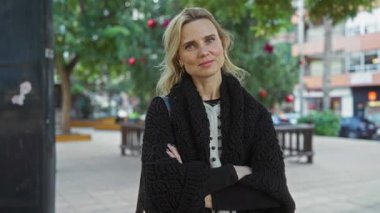 Şehir parkında kolları çaprazlanmış bir kadın, şehir merkezinde olgun güzelliğini ve sarı saçlarını sergiliyor..