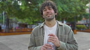 Güneşli bir günde şehir parkında elinde su şişesi tutan bir adam ağaçlar ve şehir manzarasıyla çevrili rahat bir ifadeyle..