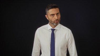 İspanyol adam, siyah arka plana karşı resmi giyinmiş, onaylanmamış bir tavır sergilerken el kol hareketi yapmaz.