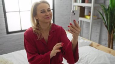 Yatak odasında akıllı telefonla sohbet eden, gülümseyen bir kadın. Pencerelerden içeri güneş ışığı süzülen kırmızı pijamalar giyiyor.