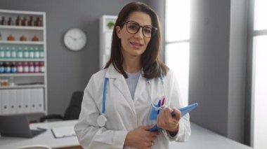 Klinikte steteskoplu kadın doktor, iş yerinde elinde tablet ve kalemle gülümsüyor..