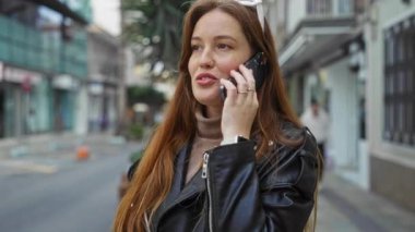 Şehir caddesinde telefonla konuşan, açık havada konuşan, kızıl saçlı, deri ceketli, şehirde güzellik ve modern yaşam tarzını sergileyen bir kadın..