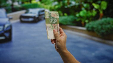Şehir sokağında elinde Hong Kong Doları banknotları olan bir adam. Arka planda arabalar ve yeşillikler var. Açık hava ve iş fırsatlarını sembolize ediyor..