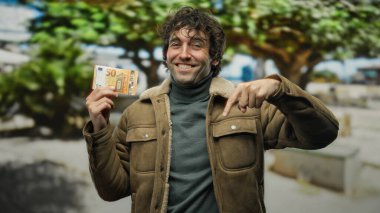 Şehirdeki kıvırcık saçlı bir adam elinde avrupa banknotunu tutarken, bronz bir ceket giyerken, sokak ortamında gülümserken, mali refah öneriyor..