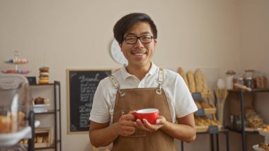 Çin 'deki bir pastanede kırmızı bardağı tutan genç adam. Etrafı fırın ve hamur işleriyle dolu. Kameraya gülümsüyor..