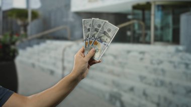 Arka planda bulanık binalar olan bir şehir caddesinde Amerikan Doları banknotları tutan adam, kentsel bir çevrede finansal temalara vurgu yapıyor.