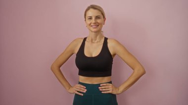 Pembe duvarın önünde kendinden emin bir şekilde gülümseyen, atletik yapısını ve günlük tarzını sergileyen, sarı saçlı, pozitiflik ve zindelik saçan bir kadın..