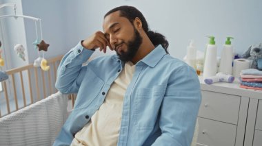 Sakallı genç adam yatak odasında uyur. Bebek beşiğinin yanında. Yumuşak yastıklarla ve yumuşatıcı pastel renklerle çevrili..