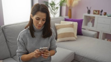 Rahat oturma odasında akıllı telefon kullanan, süveter giyen, minderler ve modern dekorlarla çevrili, ekranına odaklanmış, rahat bir iç mekan atmosferi olan bir kadın..