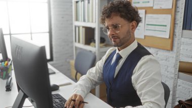 Ofiste çalışan yakışıklı bir adam, bilgisayar ekranına odaklanmış, profesyonel bir takım giymiş, dosyalar ve kırtasiyelerle çevrili, modern iş yeri estetiğini temsil eden.