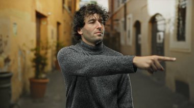 Kıvırcık saçlı genç adam şehir sokaklarını işaret ediyor. Üzerinde süveter var. Açık şehir ortamında gün boyunca kendine güvenen bir ifade sergiliyor..