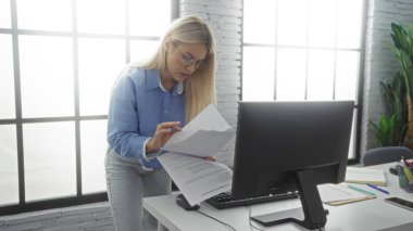 Modern ofiste çalışan gözlüklü bir kadın bilgisayarla belgeleri kontrol ediyor, profesyonelliği resmediyor ve parlak iç mekanlara odaklanıyor..