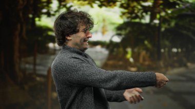 Genç bir adam parkta şakacı hareketler yaparak dingin doğal arka planla harmanlanmış gür ağaçların altında enerjik bir enerji ve karizma yayıyor.