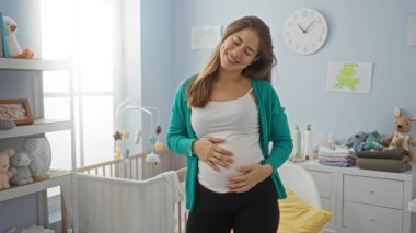 Hamile kadın yatak odasında karnını dingin bir çocuk odasında tutarken gülümsüyor. Yanında yumuşak oyuncaklar ve pastel dekorlarla çevrili bir beşik var..